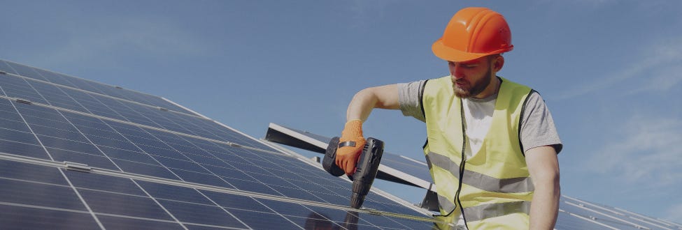 A man in an orange hard hat uses a power drill while installing solar panels on a roof.